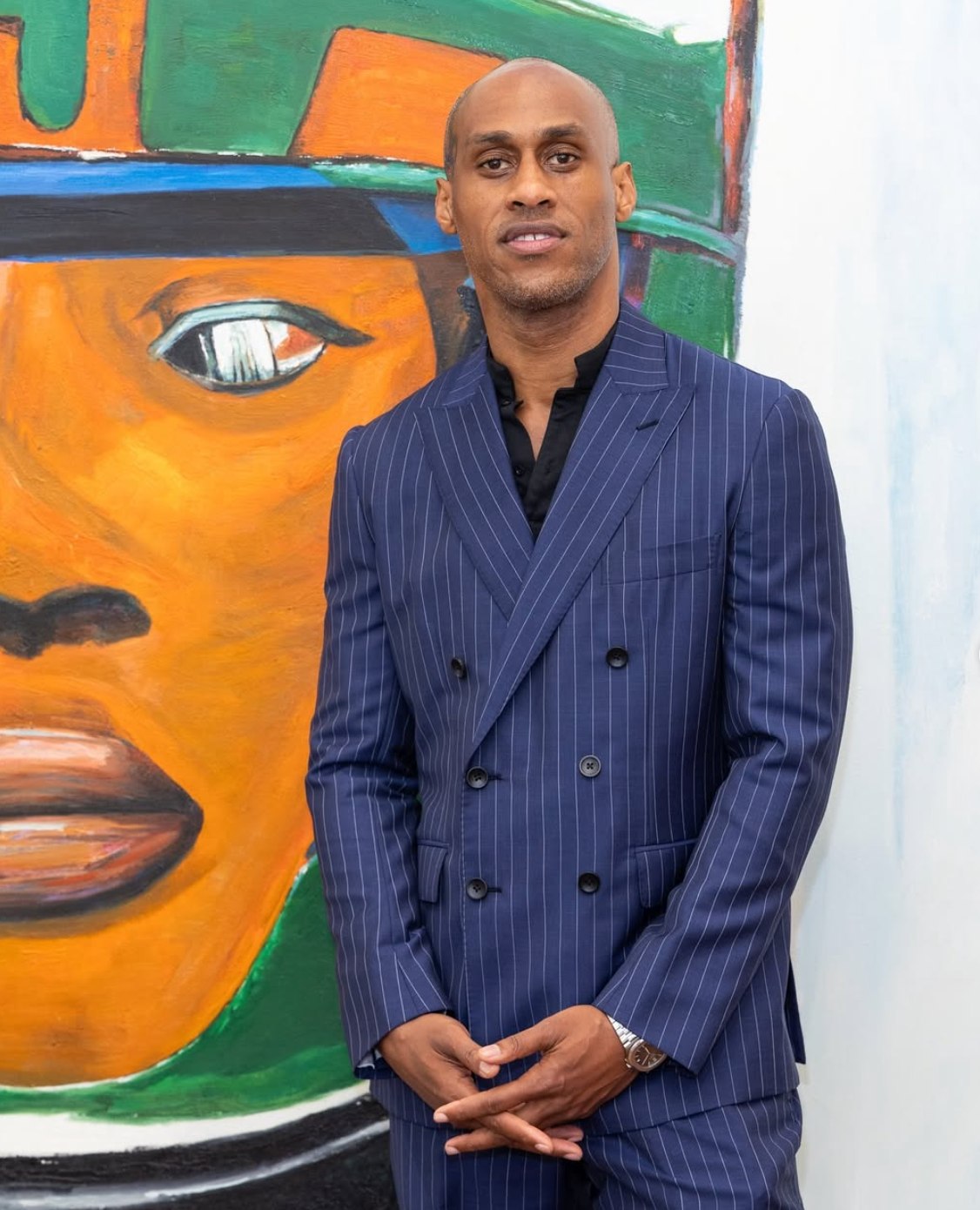Watts in a navy pinstripe double-breasted suit, standing in front of a large painted portrait in orange, green and blue